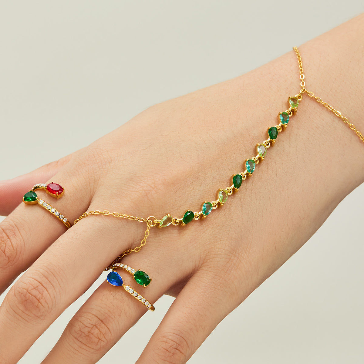 Gold-tone stainless steel hand chain bracelet with green CZ stones connecting the wrist to a finger