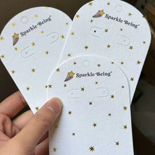 White jewelry display tags with small gold foil details held in hand