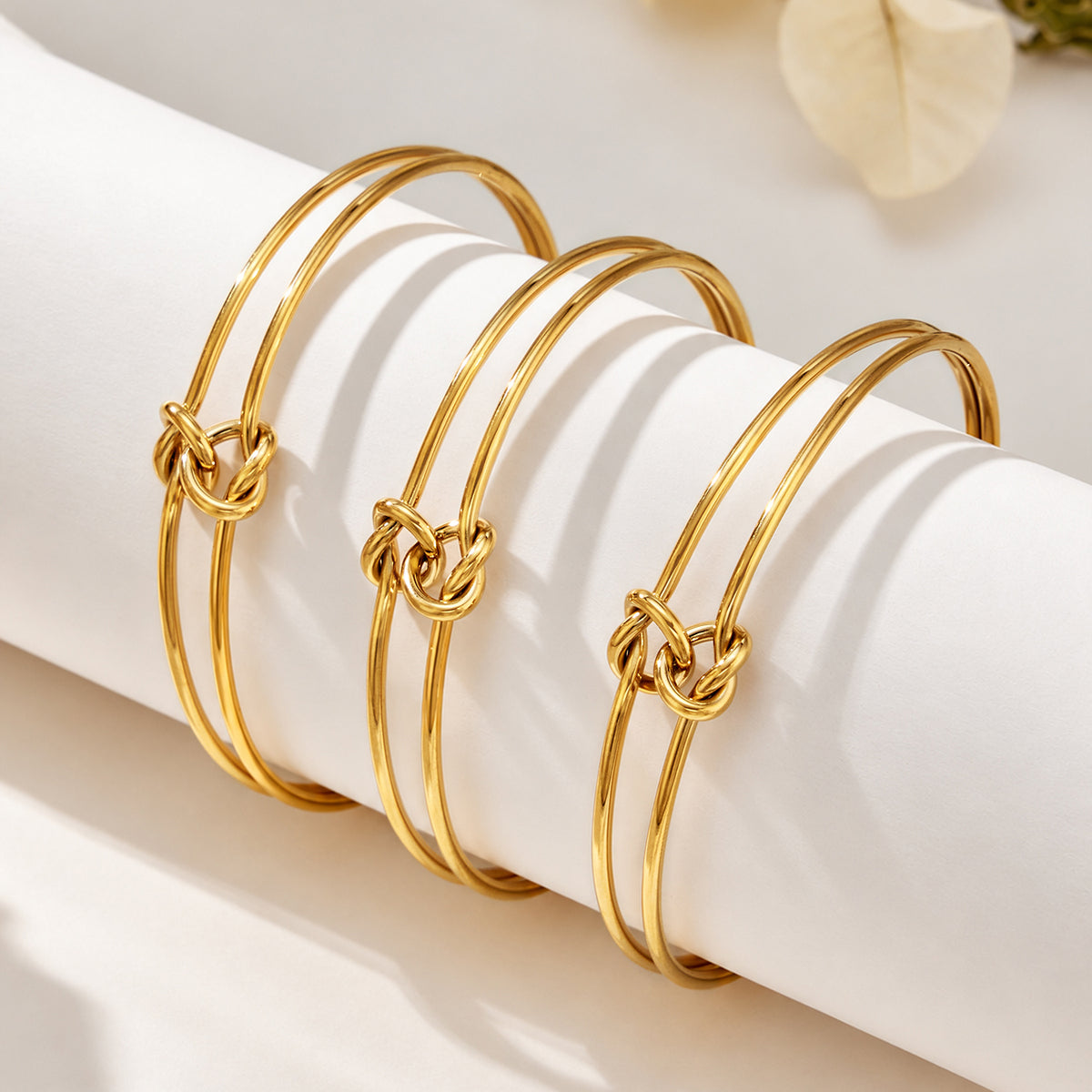 Three minimalist gold-tone knot cuff bracelets arranged on a cylindrical display