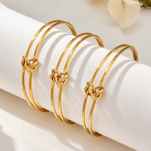 Three minimalist gold-tone knot cuff bracelets arranged on a cylindrical display