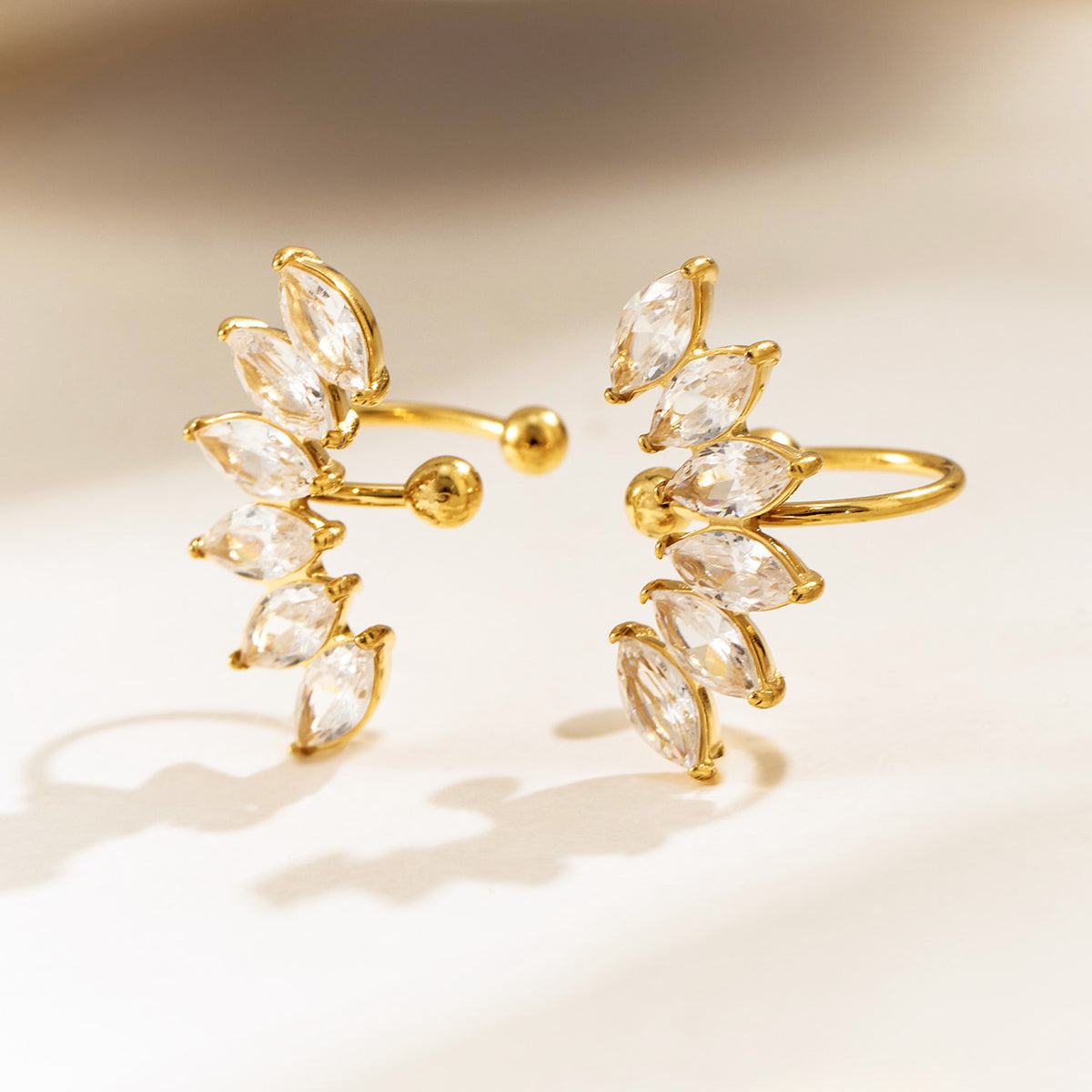 Two gold-tone leaf ear cuffs displayed on a light surface, highlighting their curved shapes and sparkling stone settings