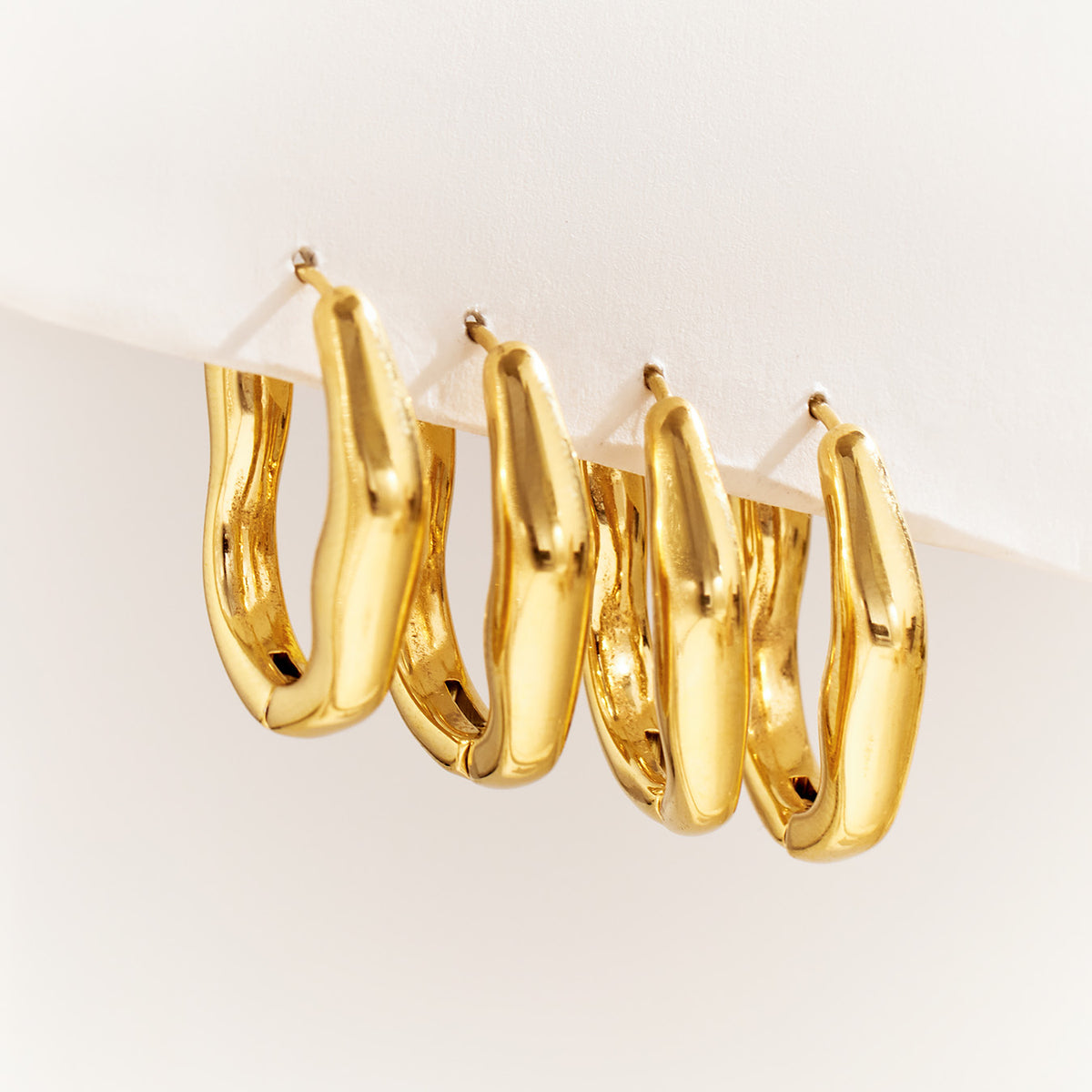 Multiple gold irregular hoop earrings displayed upright on a light surface, showing their rounded sculptural forms