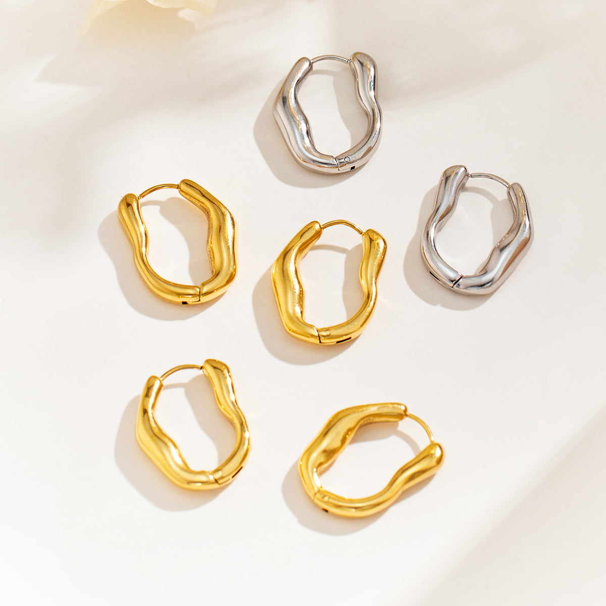 Gold and silver sculptural hoop earrings arranged on a light surface, showing contrasting polished finishes and wavy shapes