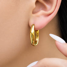 Gold-tone wavy hoop earring worn on a model's earlobe, showing the smooth metal contours and dimensional design
