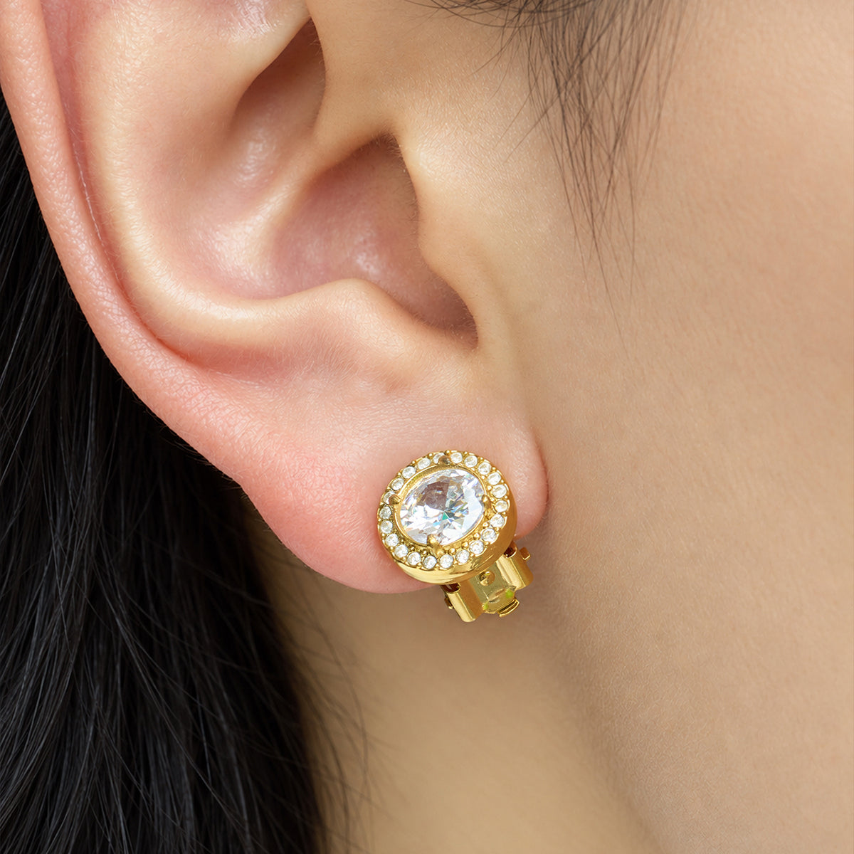 Close-up of a gold round clip-on earring on the earlobe with a large faceted clear stone surrounded by smaller sparkling stones
