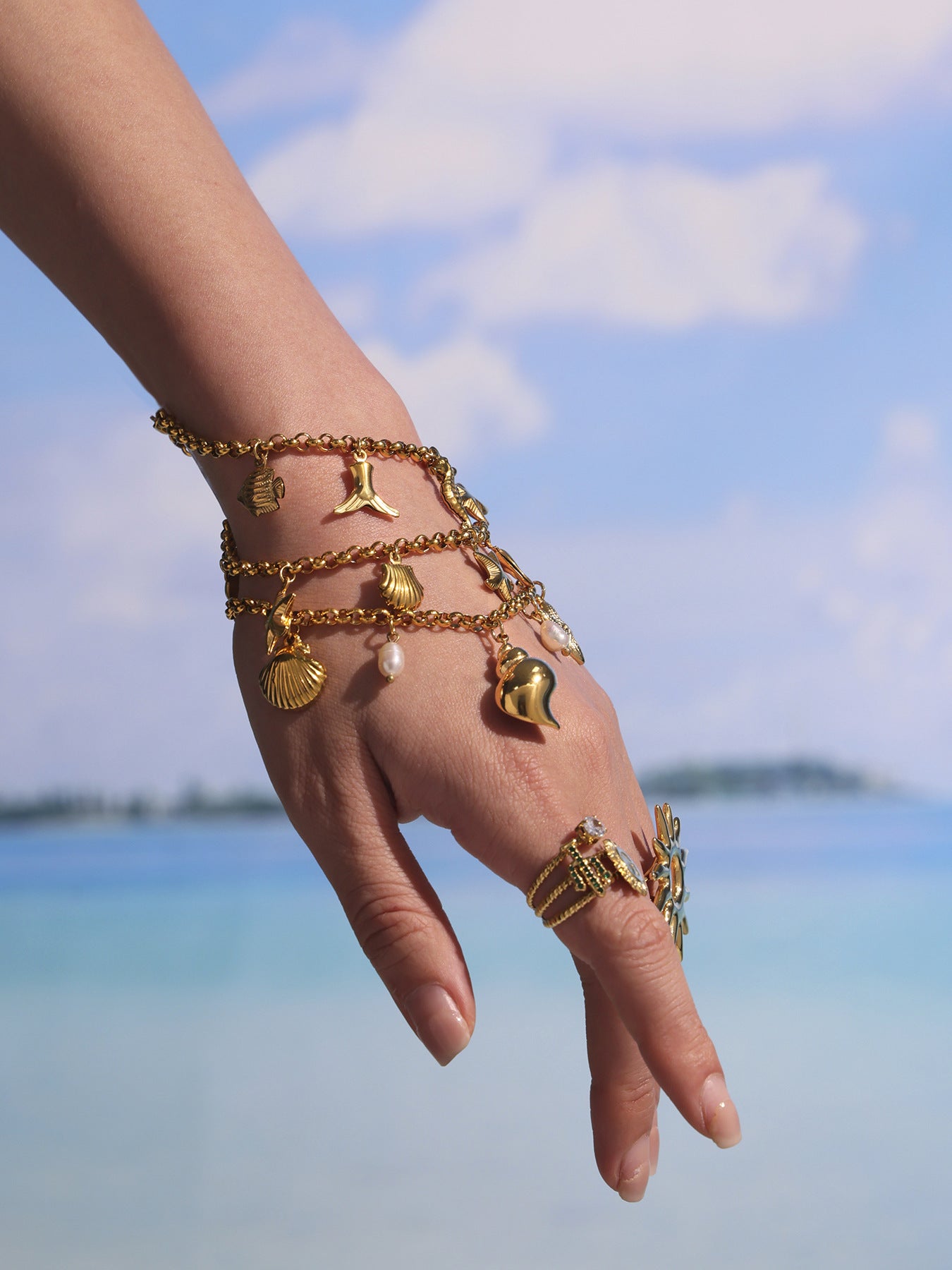 Hand wearing stacked ocean charm bracelets with shell, conch, starfish and pearl details, 18K PVD gold summer jewelry