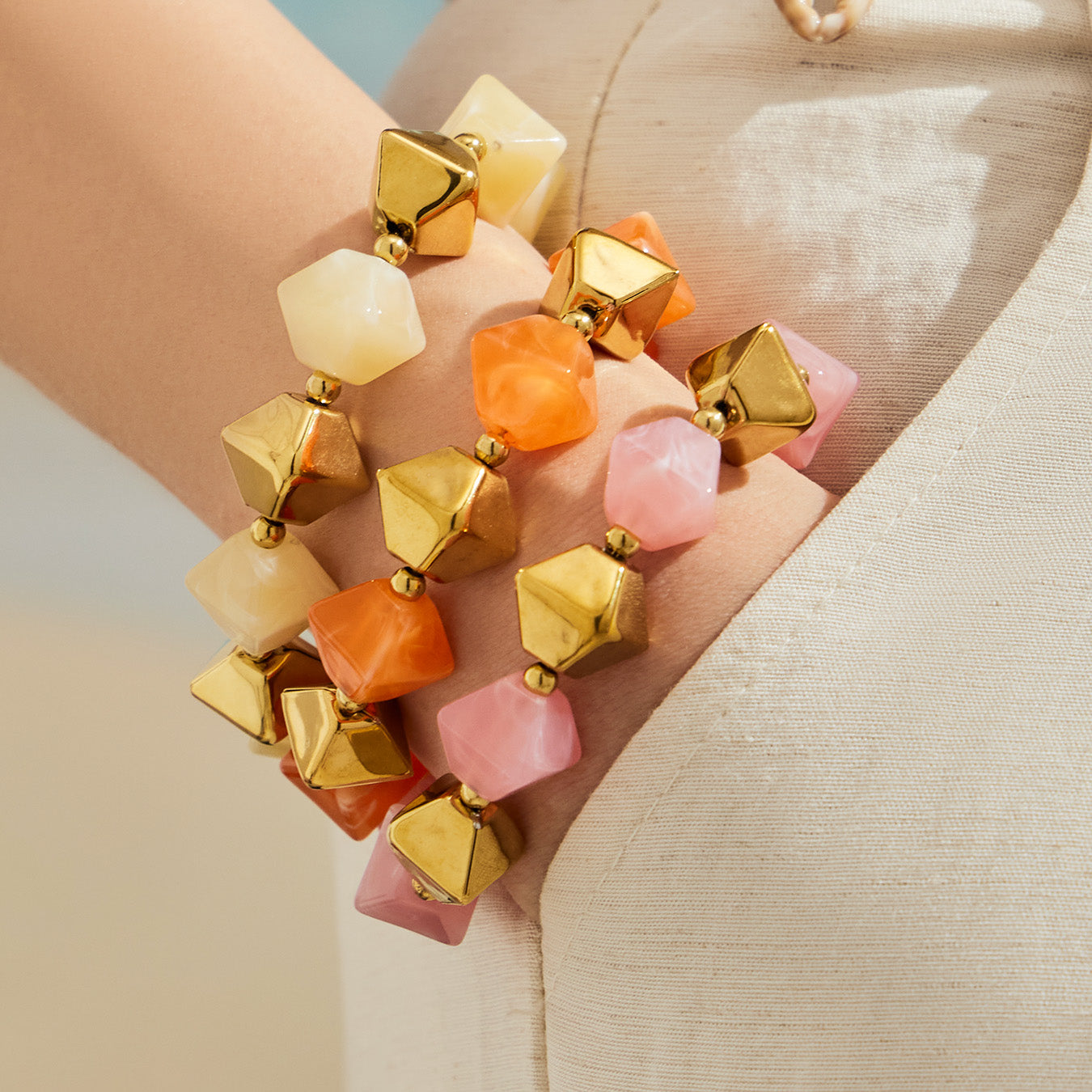 Close-up of stacked pink orange and pale yellow square resin bead bracelets with gold stainless steel accents