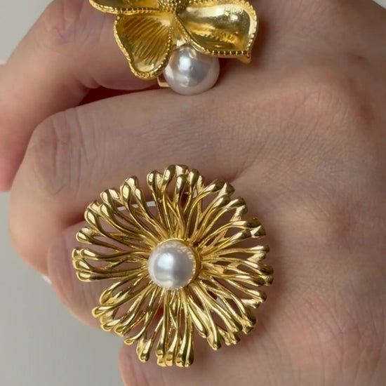 Two gold-tone statement flower rings with sculpted petals and white pearl-like accents, shown on a hand.