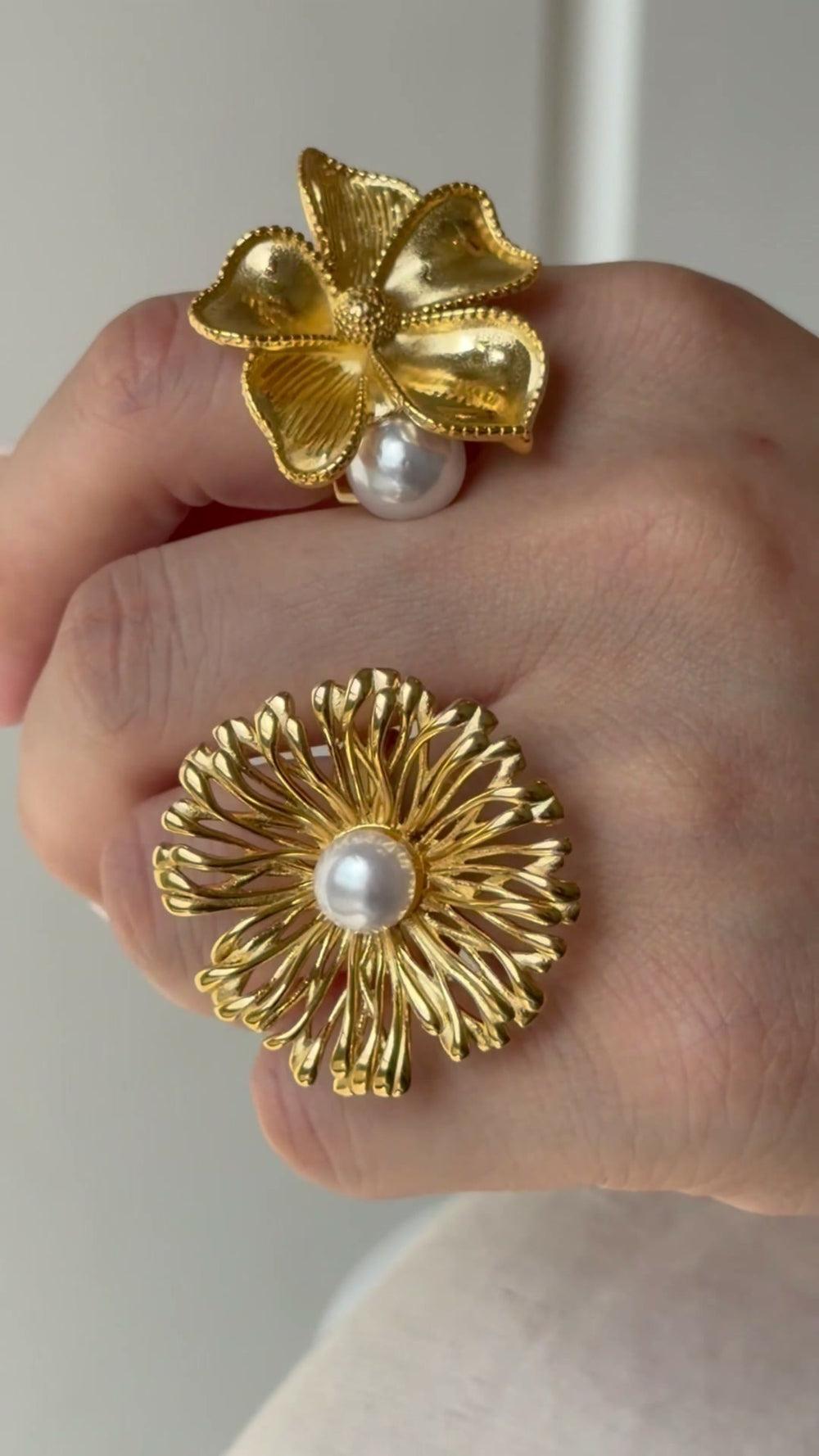 Two gold-tone statement flower rings with sculpted petals and white pearl-like accents, shown on a hand.