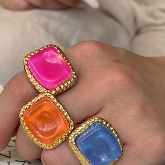 Pink, orange and blue square statement rings with textured gold-tone borders, shown on a hand.