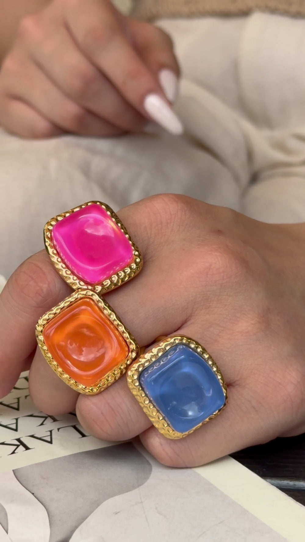 Pink, orange and blue square statement rings with textured gold-tone borders, shown on a hand.