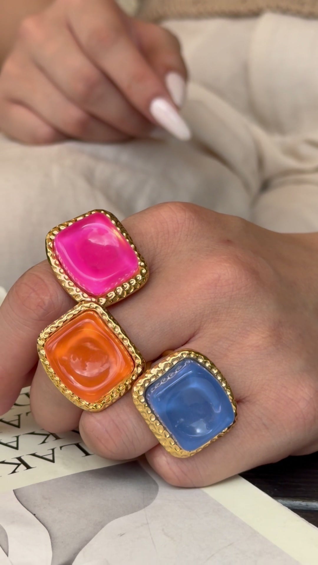 Pink, orange and blue square statement rings with textured gold-tone borders, shown on a hand.