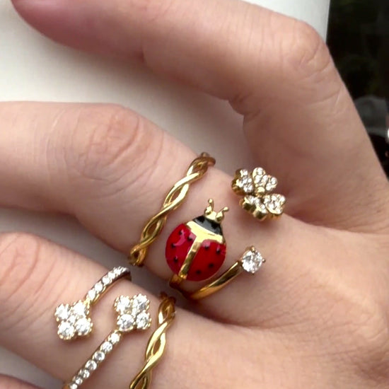 Gold-tone rings featuring a red ladybug, clear stone flowers and twisted bands, shown on a hand holding a white cup.