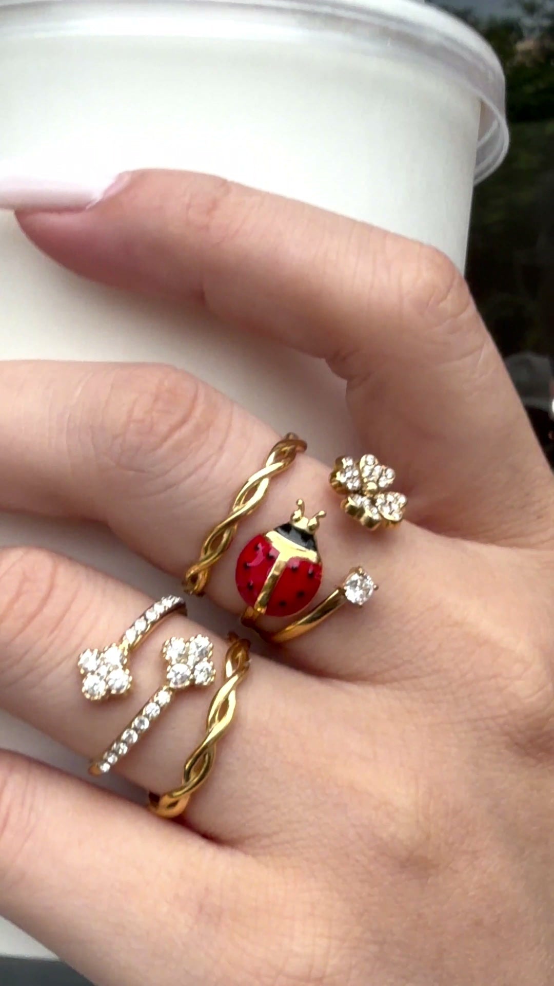 Gold-tone rings featuring a red ladybug, clear stone flowers and twisted bands, shown on a hand holding a white cup.