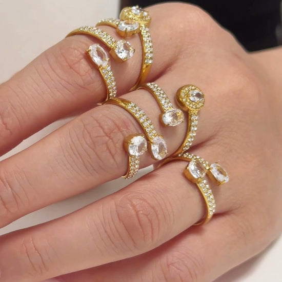 Gold-tone open rings with clear stones and curved stone-set bands, shown on a model's hand.