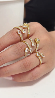 Gold-tone open rings with clear stones and curved stone-set bands, shown on a model's hand.