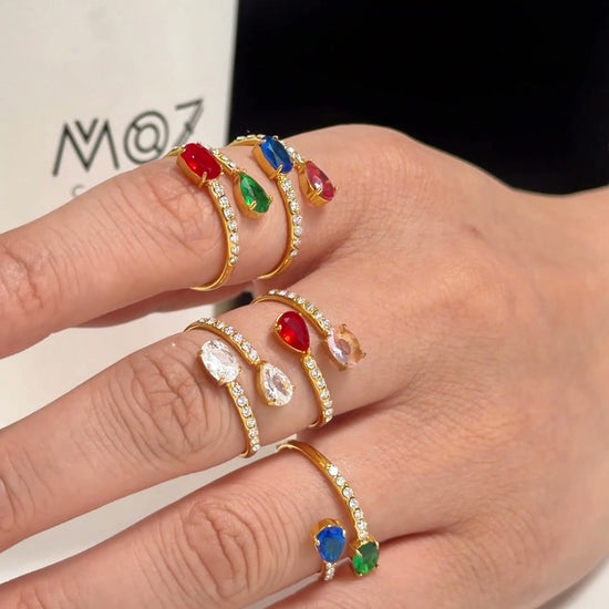 Gold-tone open rings with colorful oval stones and clear stone accents, shown on a model's hand.