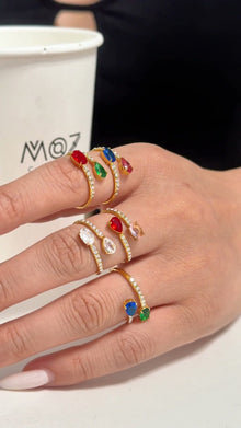 Gold-tone open rings with colorful oval stones and clear stone accents, shown on a model's hand.