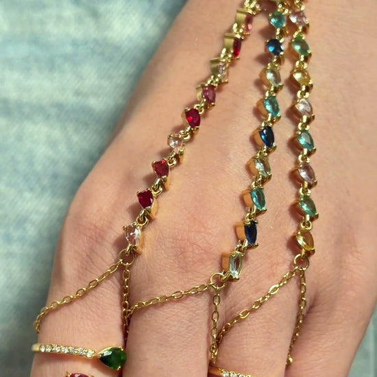 Gold-tone hand chain bracelets with colorful stones and delicate finger chains, shown on a model's hand.