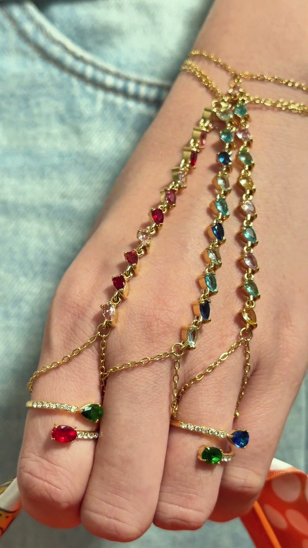 Gold-tone hand chain bracelets with colorful stones and delicate finger chains, shown on a model's hand.
