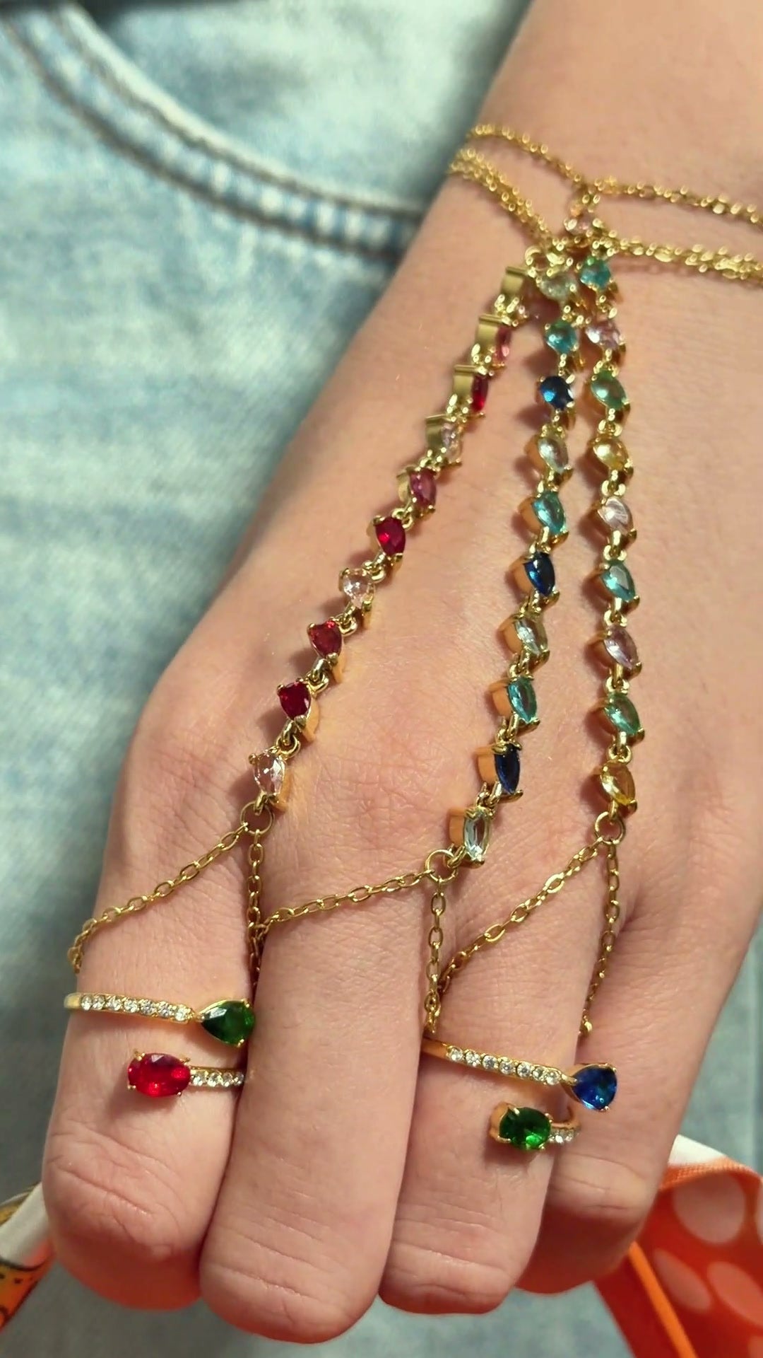Gold-tone hand chain bracelets with colorful stones and delicate finger chains, shown on a model's hand.