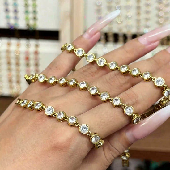 Gold-tone tennis bracelet with round clear stones in bezel-style settings, shown by hand for wholesale jewelry buyers.