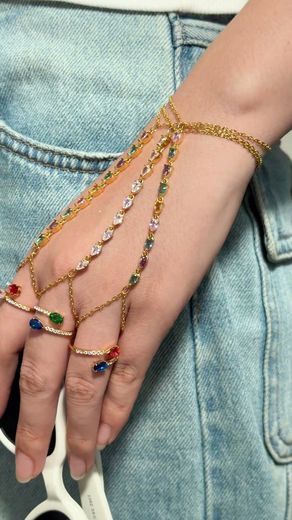 Gold-tone hand chain jewelry with multicolored stones and delicate chains connecting the wrist to the fingers.