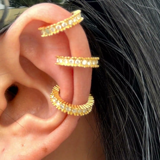 Gold-tone ear cuffs with clear stone accents and textured edges, shown stacked on a model's ear.