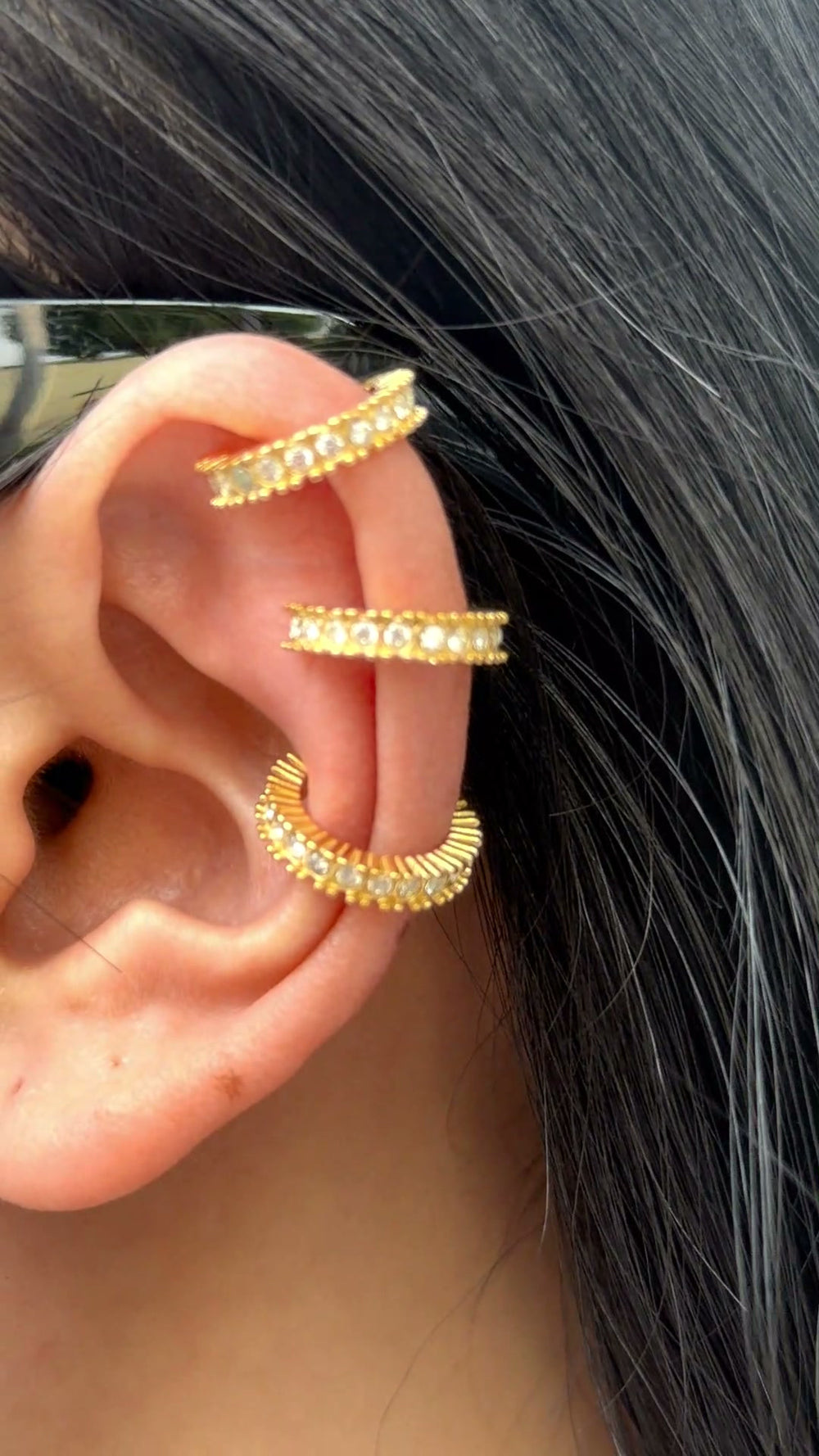 Gold-tone ear cuffs with clear stone accents and textured edges, shown stacked on a model's ear.