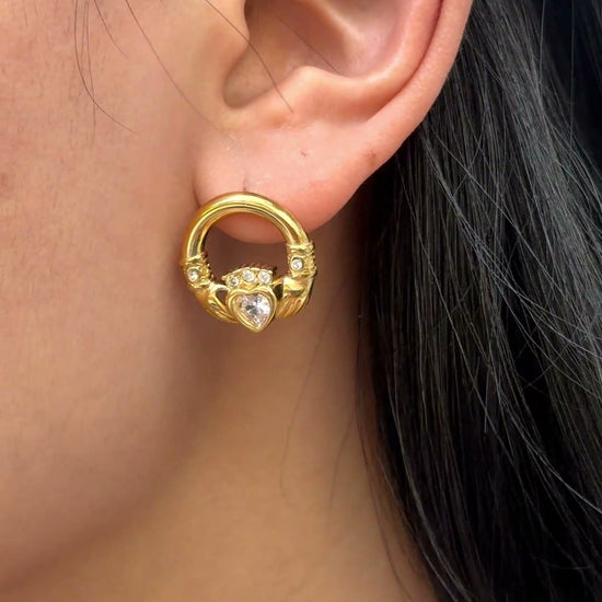Gold-tone circular earrings with a heart-shaped clear stone and decorative details, shown on a model.