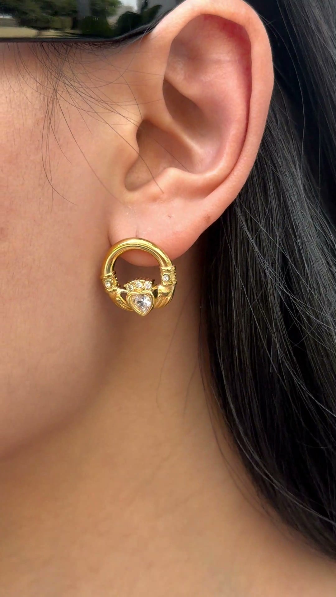 Gold-tone circular earrings with a heart-shaped clear stone and decorative details, shown on a model.