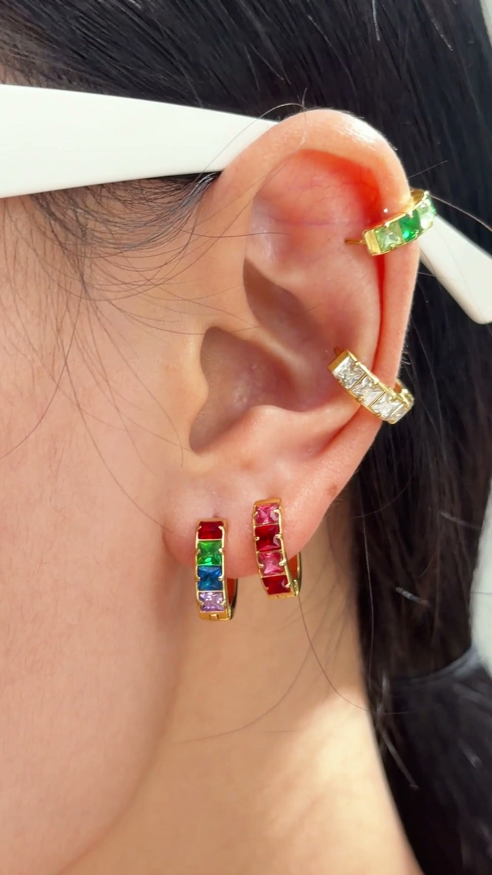 Gold-tone hoop earrings with colorful rectangular stones in rainbow and pink tones, shown on a model's ear.