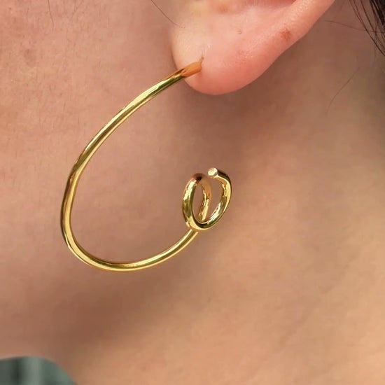 Gold-tone open hoop earrings with curled ends, shown on a model.