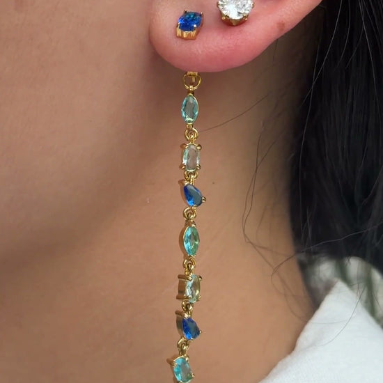 Long gold-tone earrings with blue and aqua stones in mixed shapes, shown on a model.
