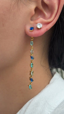 Long gold-tone earrings with blue and aqua stones in mixed shapes, shown on a model.