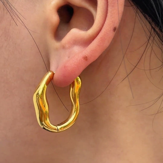 Polished gold-tone hoop earrings with an irregular wavy outline, shown on a model's ear.