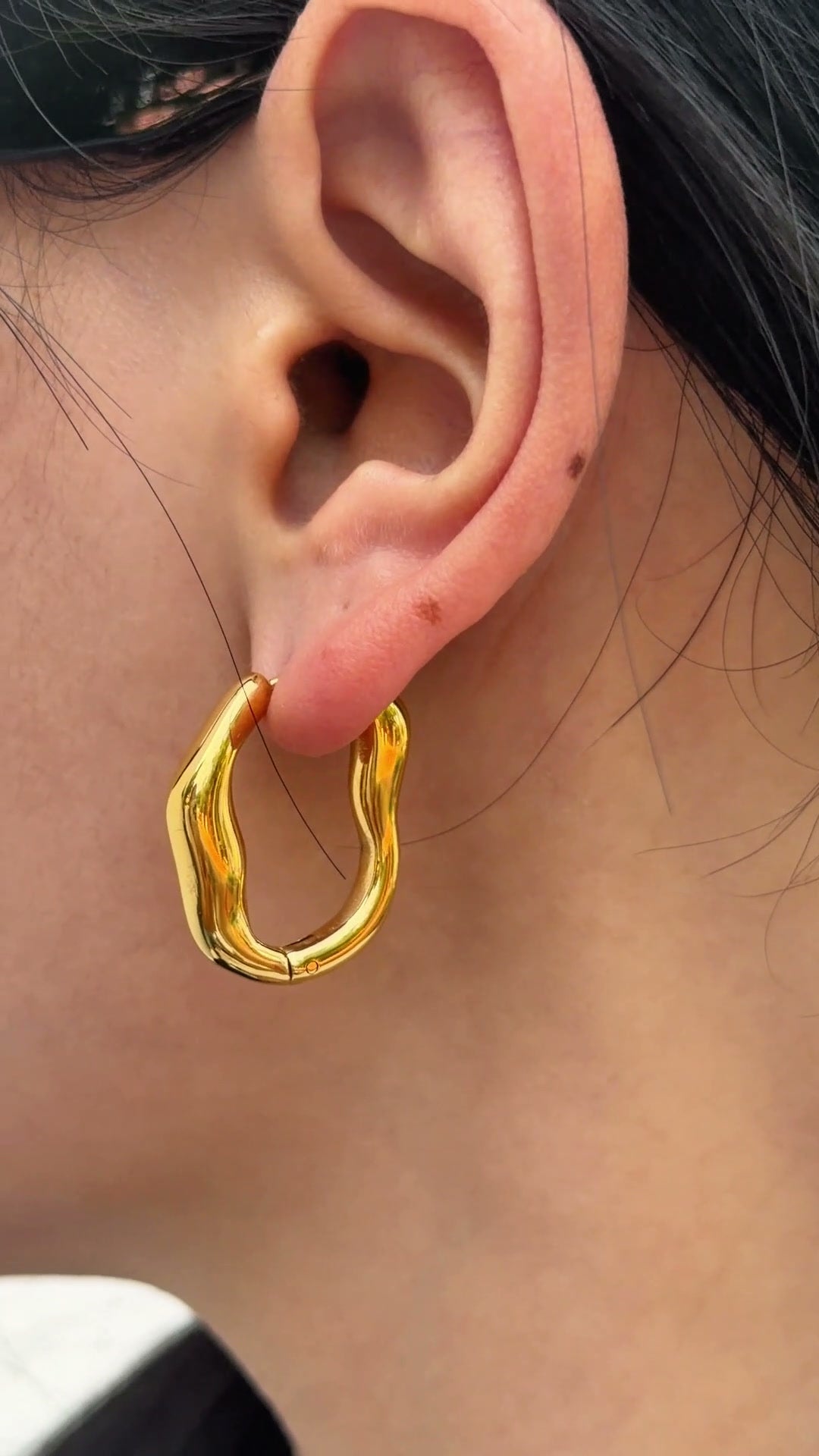 Polished gold-tone hoop earrings with an irregular wavy outline, shown on a model's ear.