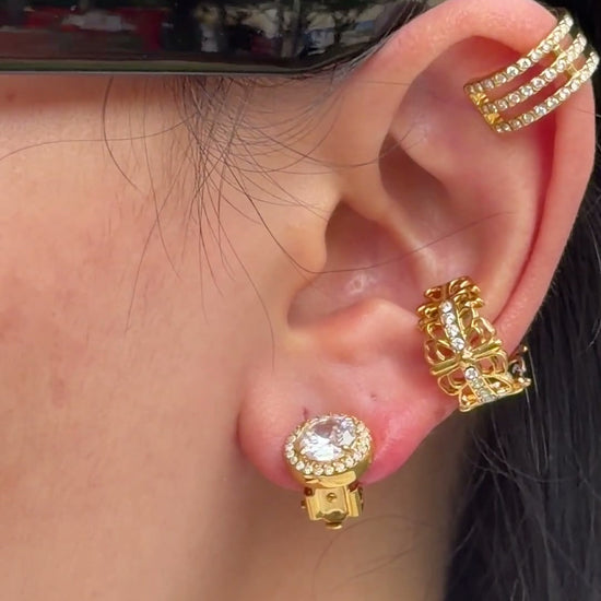 Gold-tone earrings featuring a round clear center stone surrounded by a halo of smaller stones, shown on a model.