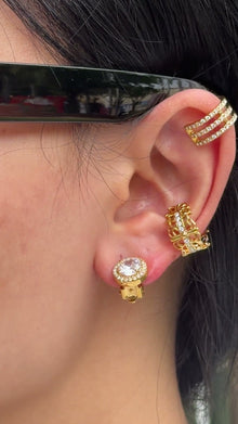 Gold-tone earrings featuring a round clear center stone surrounded by a halo of smaller stones, shown on a model.