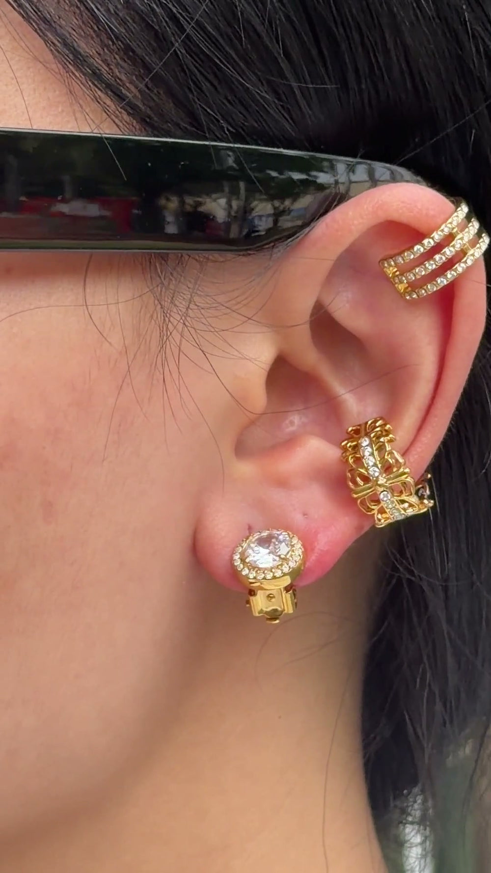 Gold-tone earrings featuring a round clear center stone surrounded by a halo of smaller stones, shown on a model.