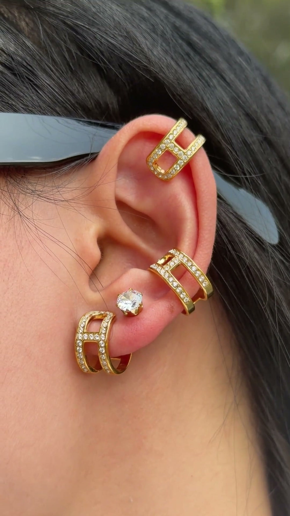 Gold-tone geometric hoop earrings with open rectangular details and clear stone accents, shown on a model's ear.