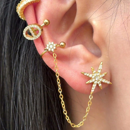 Gold-tone starburst stud and floral ear cuff connected by a chain, with clear stone accents, shown on a model.