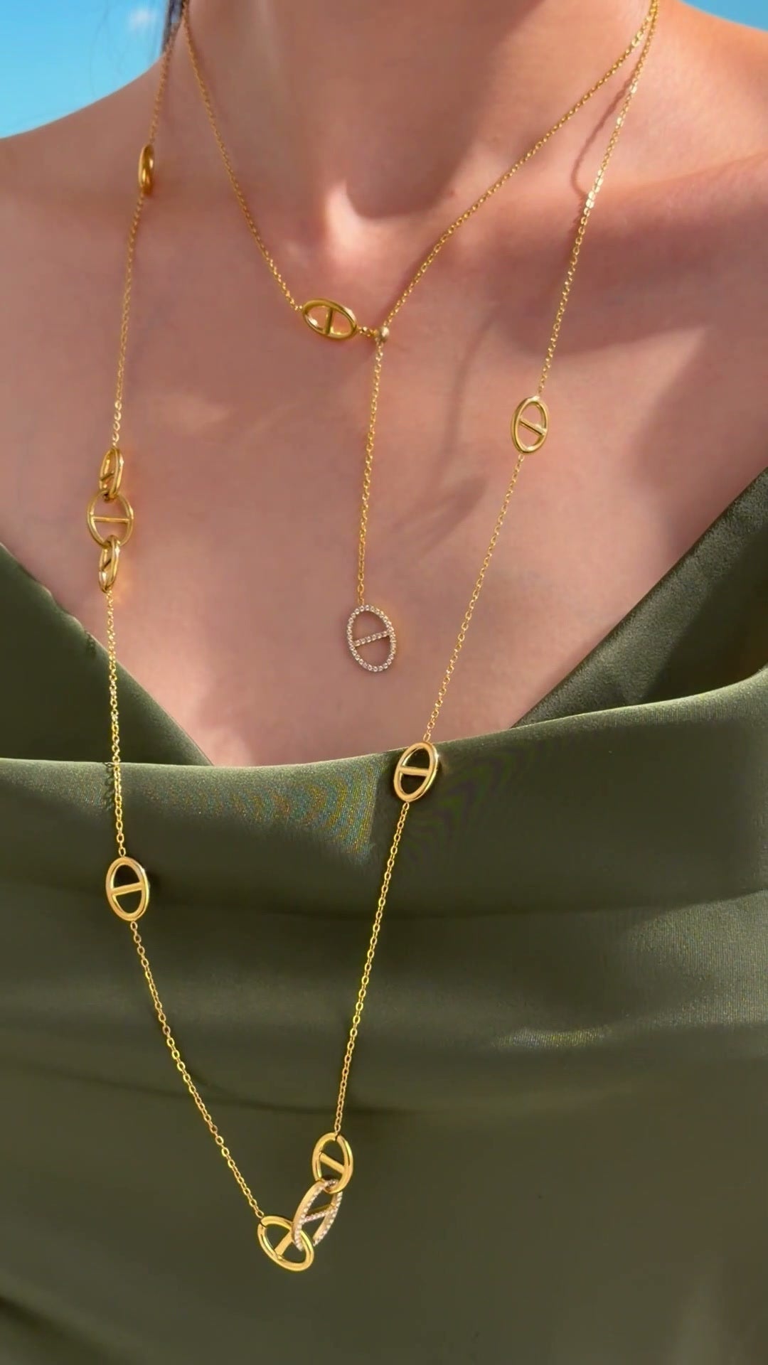 Layered gold-tone necklaces with oval mariner-link stations and clear-stone accents, styled with an olive-green satin top.