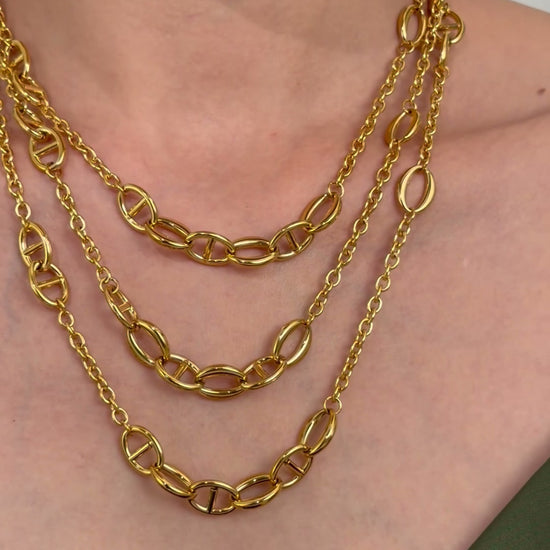 Three layered gold-tone necklaces with polished oval and mariner links, styled on a model wearing an olive-green top.
