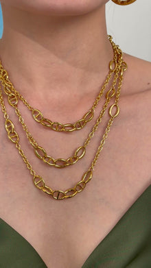 Three layered gold-tone necklaces with polished oval and mariner links, styled on a model wearing an olive-green top.