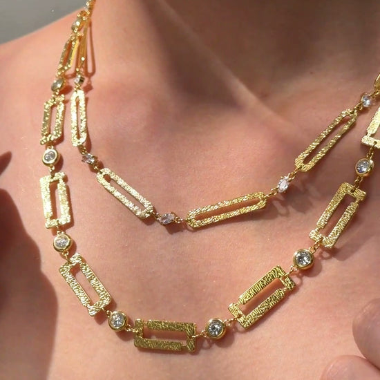 Layered gold-tone necklaces with textured rectangular links and clear sparkling accents shown on a model.