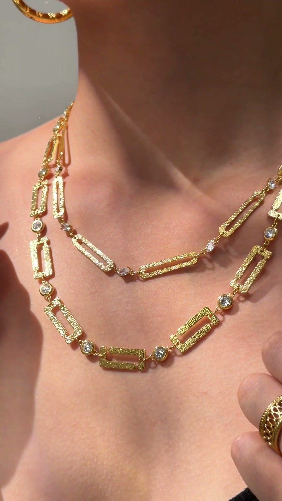 Layered gold-tone necklaces with textured rectangular links and clear sparkling accents shown on a model.