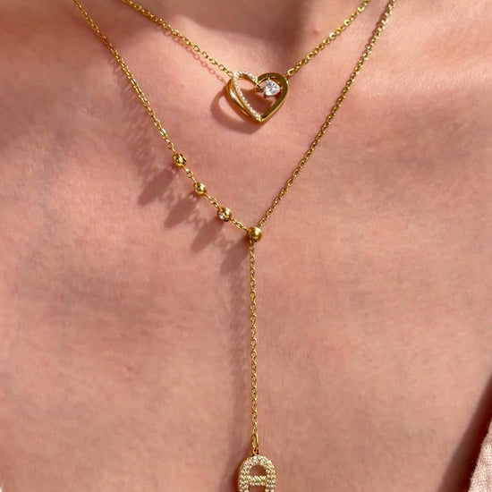 Layered gold-tone necklaces featuring an open-heart pendant and a beaded lariat with a clear-stone oval pendant, shown on a model.