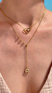 Layered gold-tone necklaces featuring an open-heart pendant and a beaded lariat with a clear-stone oval pendant, shown on a model.