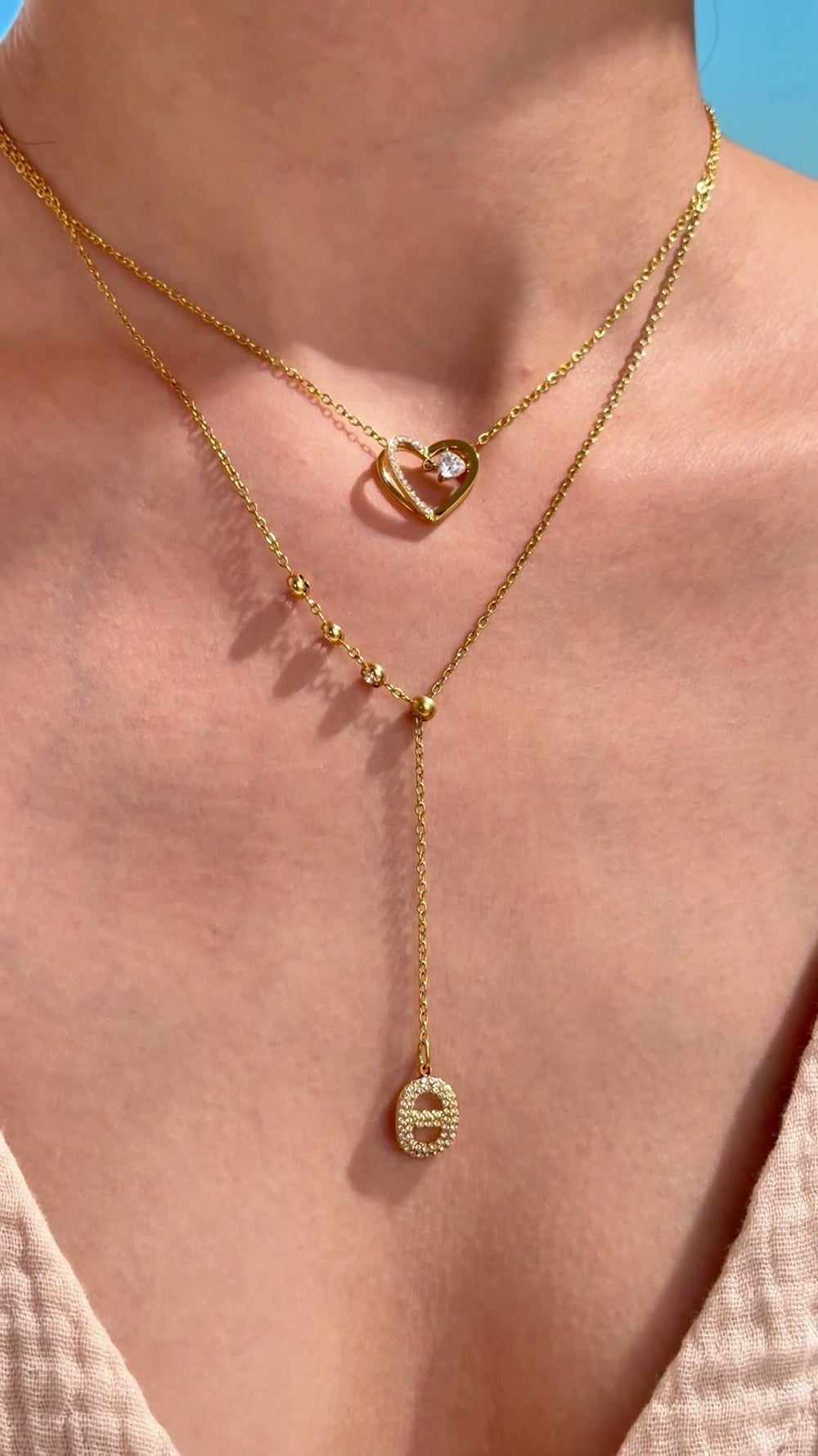 Layered gold-tone necklaces featuring an open-heart pendant and a beaded lariat with a clear-stone oval pendant, shown on a model.